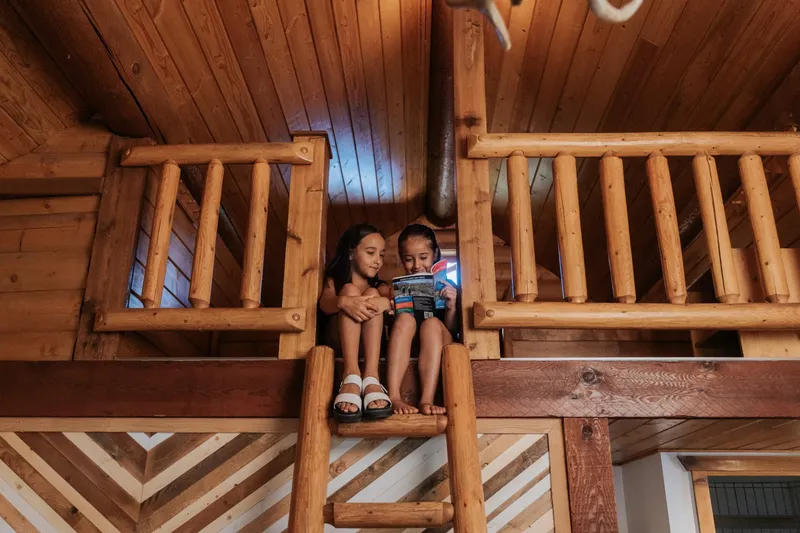 Two children sit on a wooden loft railing inside Baker Creek's log cabin, surrounded by natural timber beams.