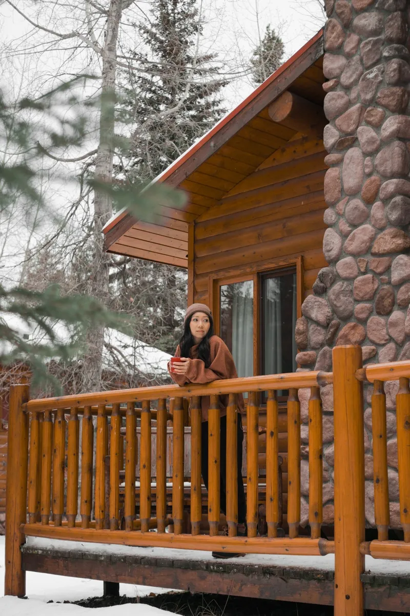 Snow-dusted log cabin at Baker Creek with stone chimney and wooden deck among evergreens.