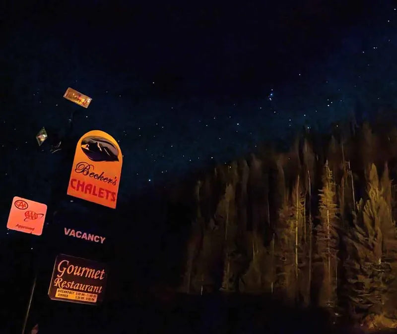Neon signs for Becker's Chalets and Gourmet Restaurant glow against dark forest at night.