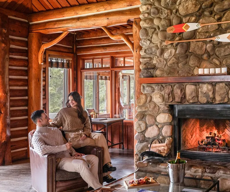 Fairmont Jasper Park Lodge lounge with log timber beams, stone fireplace, and guests by tall windows.