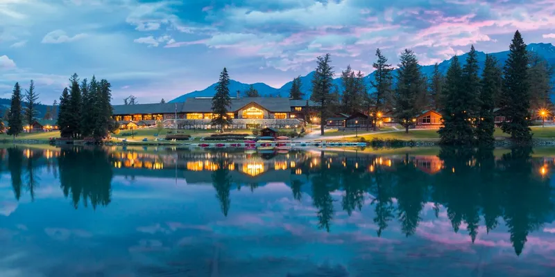 Fairmont Jasper Park Lodge's illuminated log buildings and cabins reflect in the lake at dusk.