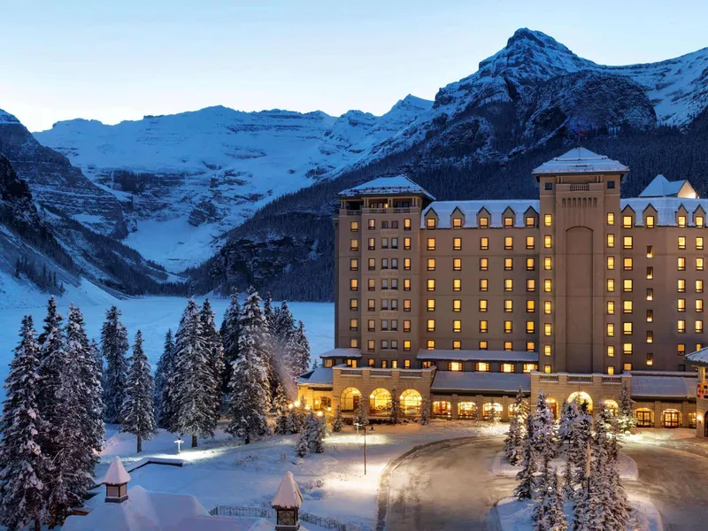 Fairmont Chateau Lake Louise glows at dusk beside frozen lake and snow-covered peaks.