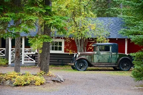 Log cabin with red walls and white trim beside weathered vintage truck amid tall pines at Johnston Canyon Lodge.