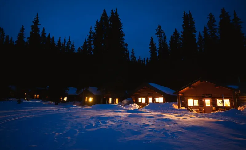 Log cabins at Shadow Lake Lodge glow warmly at dusk against deep snow and forested peaks in winter.