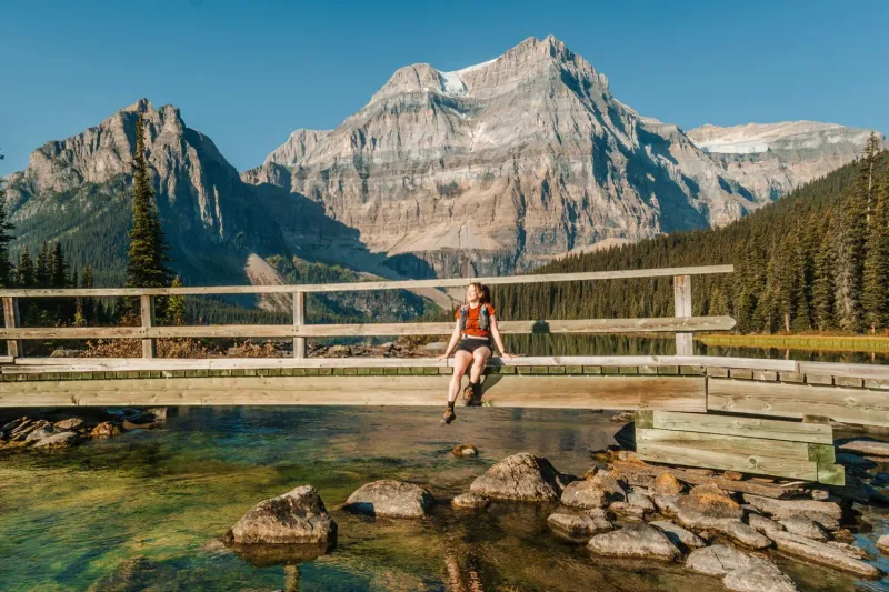 Hiker stands on Shadow Lake Lodge's wooden deck overlooking turquoise alpine lake and layered mountain peaks.