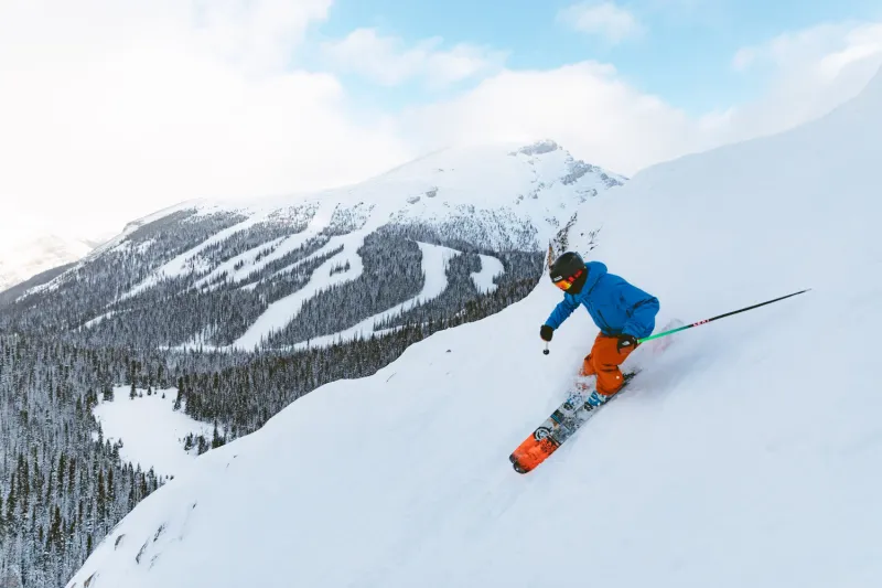 Skier descending snowy slope at Sunshine Mountain Lodge with snow-covered peaks and forested ridges in background.