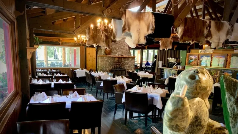 Sunwapta Falls Lodge dining room with timber beams, stone fireplace, hides overhead, and mountain views through windows.